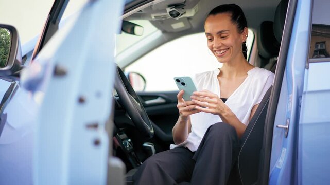 Young cheerful Hispanic woman using a smartphone while sitting in the driver&rsquo;s seat of a parked car, capturing modern lifestyle and mobile communication.