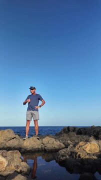 Contemplative mature man standing on rugged coastal rocks, looking at the ocean and his reflection in a tide pool on a sunny day with clear blue sky, enjoying a moment of peaceful solitude