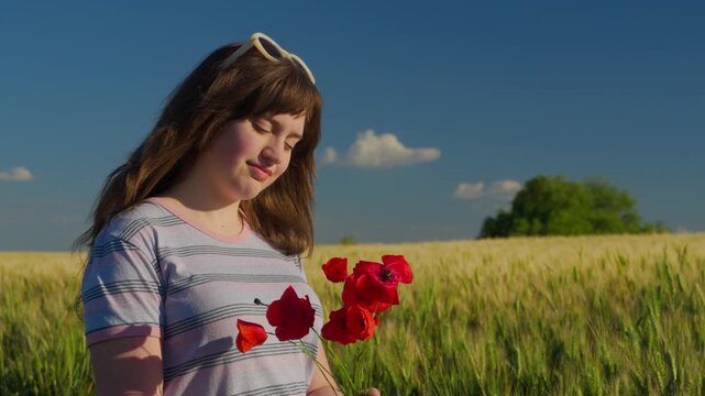 A beautiful young teenager smiling peacefully while holding vibrant red poppy flowers in a vast wheat field under a clear blue sky at sunset