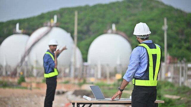 Engineers wearing safety gear, including hard hats examining survey a large blueprint tablet standing industrial facility gas or oil refinery engaged in a job requires high safety standards concept.	