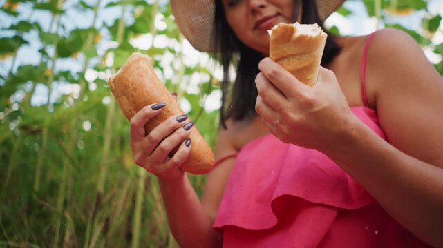 Close up of woman in pink holding and breaking bread with green plants blurred in background. Hands grasp baguette soft lighting highlights texture details. Outdoor setting with natural