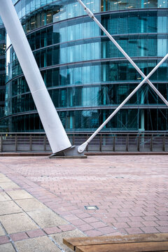 White structural columns and diagonal braces frame a curved glass office facade beside a paved urban plaza in a modern architectural scene