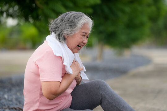 Senior woman chest pain cardiac emergency outdoors towel, seated on path clutching chest, breathing hard, possible heart problem, urgent medical concept