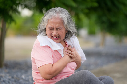 Senior woman chest pain heart attack emergency outdoor park exercise, sitting on ground with towel, clutching chest, distressed face, urgent medical problem after workout