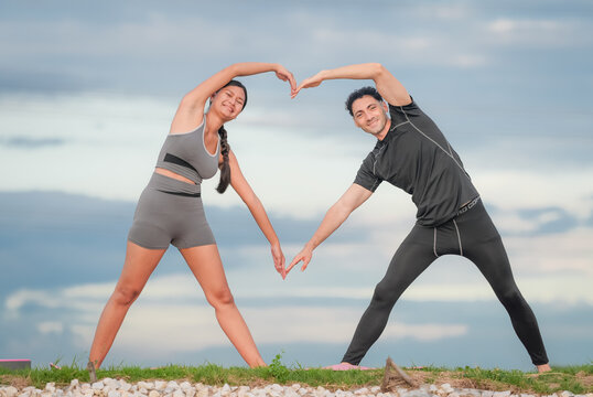 Couple stretching fitness yoga workout outdoor heart flexibility pose on grass under cloudy sky, smiling partners in activewear practicing side bend exercise, calm wellness mood