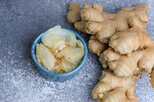 Pickled ginger served in a small bowl next to fresh ginger roots on a gray surface