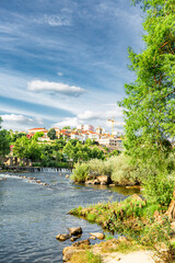 Fototapeta premium Amarante, Douro Valley, Portugal Amarante town rises above a winding river, framed by lush greenery. Blue sky with scattered white clouds adds to the serene atmosphere. Water flows gently over rocks, 