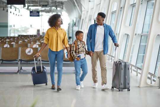 A smiling African American family with a young son walks hand-in-hand through a modern airport terminal, pulling their suitcases and ready for travel