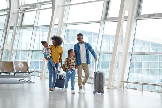 A smiling African American family with two children is traveling together, pulling suitcases and holding boarding passes in a modern airport concourse