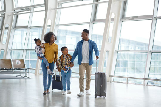 A smiling African American family walks together through a modern airport terminal, pulling suitcases and holding travel documents