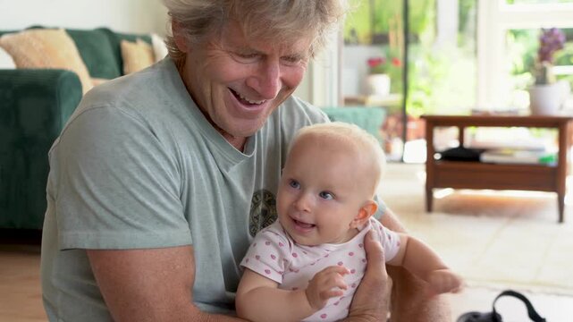 Happy grandfather playing with his baby granddaughter at home in a bright living room.