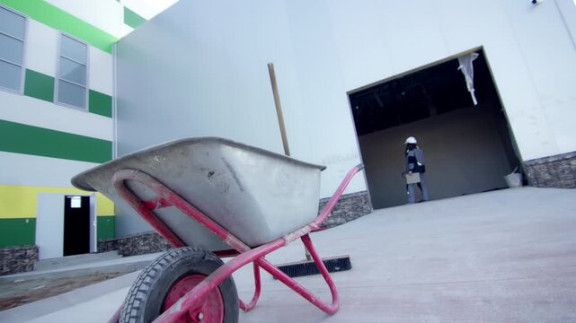 Active commercial site entrance with wheelbarrow placed in close foreground. Worker stands in high-ceiling bay doorway framed by paneling