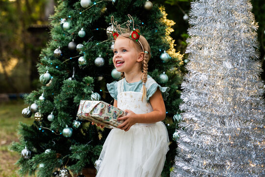 Joyful girl holding a wrapped gift at Christmastime outdoors by a silver tinsel tree