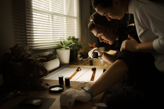 Multiethnic couple examining analog film strips on light table in darkroom. Image represents photography education and creative hobby marketing, medium shot