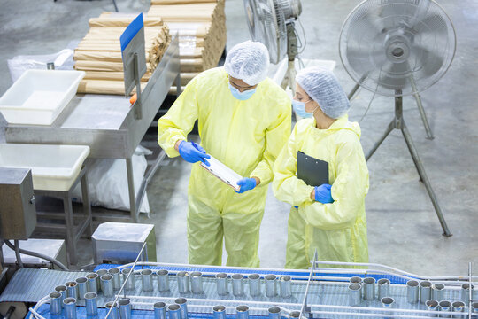 Food factory workers in yellow protective suit and hairnet checking production line with clipboard, Quality control inspectors in safety gear reviewing checklist at canned goods manufacturing line
