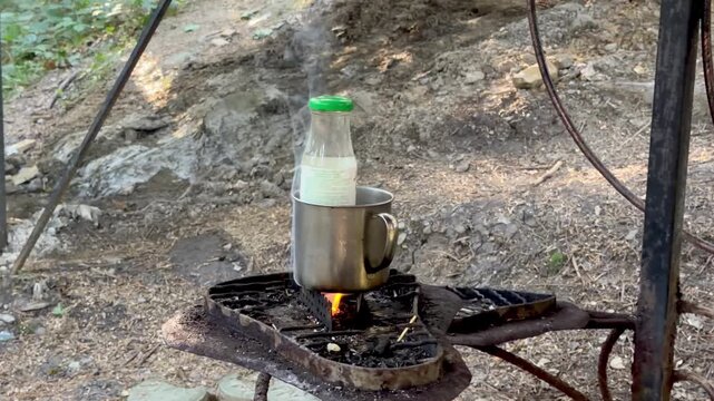Glass bottle with mineral water heating in metal cup over small campfire outdoors