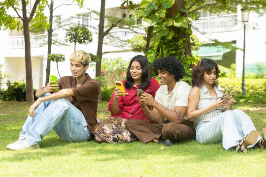 Group of indian Gen Z young adults friends busy using smartphone. while sitting in summer park, concepts of social media addiction. Digital communication. Youth culture. Wireless technology.