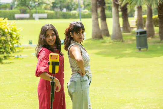 Two smiling young indian Gen Z girl or women recording video content for social media using a smartphone while standing together in summer park, Wireless Technology. Youth Culture.