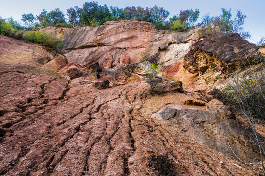 Eroded red bauxite terrain leading toward a colorful exposed rock wall in a former mine, creating strong natural lines, textures and geological patterns beneath surrounding vegetation.