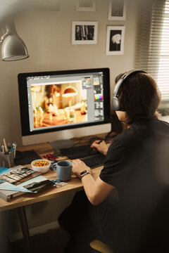Rear view of young Asian man wearing headphones, editing photo on computer using graphics tablet at home office. Image suitable for remote work and digital creator campaigns, vertical
