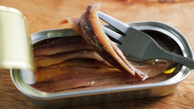 Canned anchovy fillets in oil inside an open metal tin with a fork lifting one fillet on a wooden table.