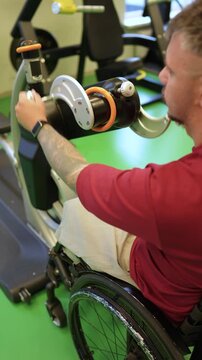 Disabled man in wheelchair exercising on hand cycling fitness machine in gym at rehabilitation session, male athlete with injury spinning ergometer with hands, increasing upper-body muscle endurance.