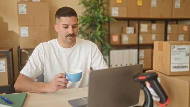 Man holding a mug while working on a laptop and checking packages in a small business building; quiet concentration.