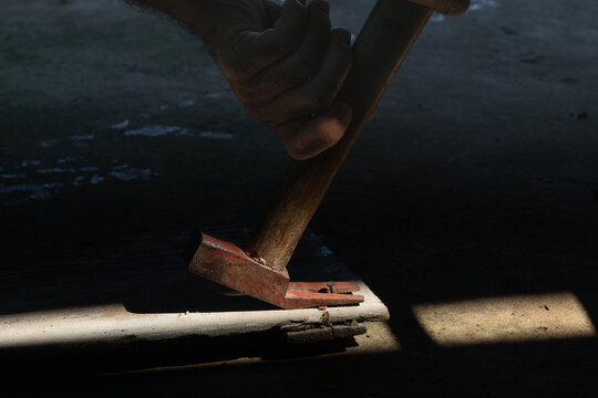  Hands of a craftsman using a claw hammer to pull out an old rusty nail from wood in moody lighting
