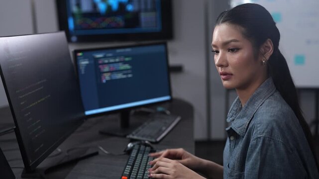 Asian female software developer coding on multiple monitors in a dark office. Smiling woman programmer working on IT systems. Corporate technology diversity and cybersecurity data development concept