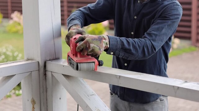 A worker uses a power sander to smooth a wooden fence outdoors during home renovation and maintenance work.