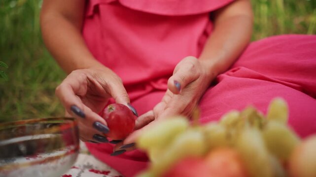 Close up of woman in pink clothing holding and examining red fruit with dark nail polish surrounded by fresh produce like grapes. scene features soft natural lighting and outdoor setting