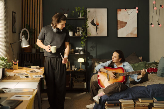 Asian young man holding mug and talking to Caucasian young woman playing acoustic guitar in bedroom, representing creative collaboration and youth lifestyle marketing