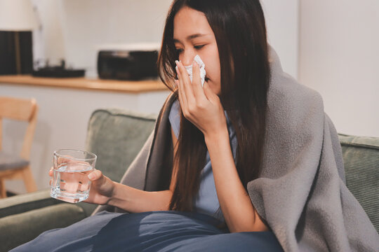 sick asian young woman sneezing into tissue paper while covered with a blanket and sitting on sofa in the living room
