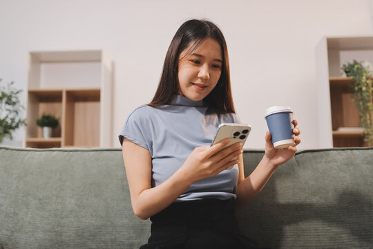 Freelance Asian woman holding mobile phone typing on laptop and working from home. Happy young girl sitting on sofa in living room.