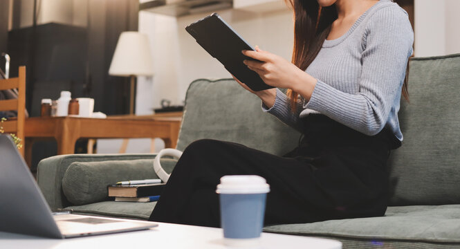 Freelance Asian woman holding mobile phone typing on laptop and working from home. Happy young girl sitting on sofa in living room.