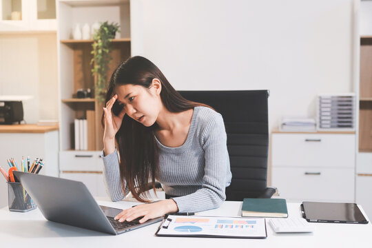 Young Asian woman sits at office desk rubbings her eyes and temples feeling pain. Stress. And exhaustion from long hours working on the computer