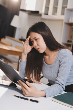 Young Asian woman sits at office desk rubbings her eyes and temples feeling pain. Stress. And exhaustion from long hours working on the computer