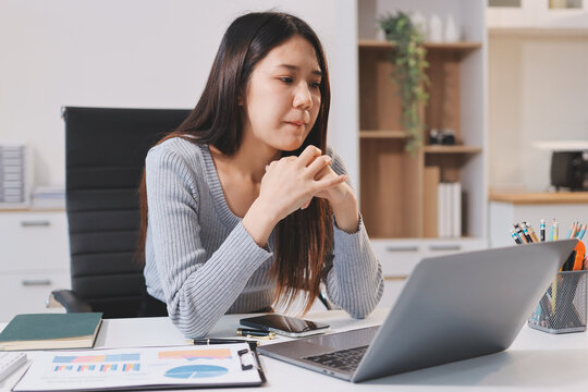 Young Asian woman sits at office desk rubbings her eyes and temples feeling pain. Stress. And exhaustion from long hours working on the computer