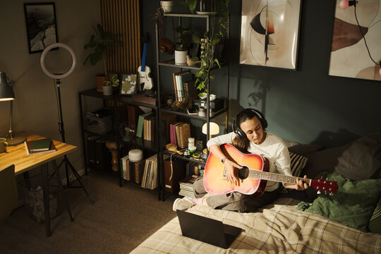 Young Caucasian woman wearing headphones playing acoustic guitar in bedroom while looking at laptop. Image ideal for online music lessons or creative hobby marketing, high angle view