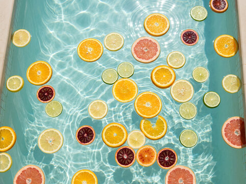 A highly realistic top-down food and lifestyle photograph of colorful citrus slices floating in a shallow glass-like layer of clear water together with a few fresh green herb leaves.