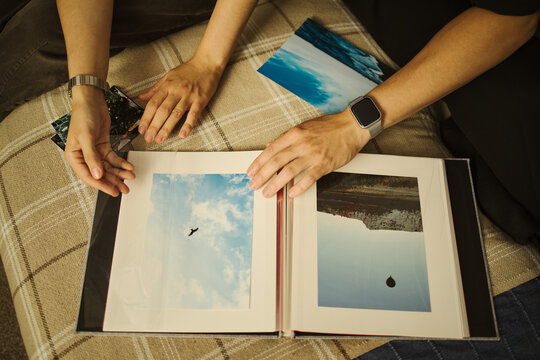 Hands of couple organizing printed travel photos in album, preserving memories. Concept of nostalgia and family lifestyle, high angle view