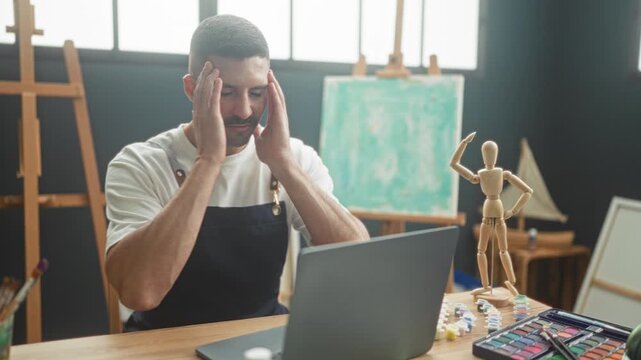 Man with hands to face at laptop in art studio with easel, canvas, paints and wooden mannequin; creative frustration.