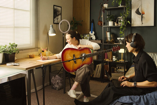 Caucasian young woman holding acoustic guitar and writing lyrics while Caucasian young man uses laptop in home studio, representing creative collaboration and music production