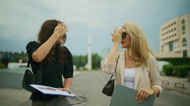 Two professional businesswomen are engaged in a discussion outdoors, holding documents and clipboards. They are standing and conversing, likely about work-related matters in an urban setting.