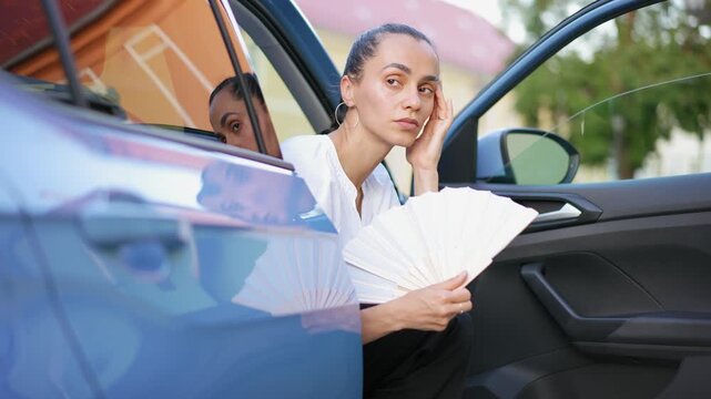 Tired Hispanic woman fanning herself while sitting in a parked car during unbearable summer heat in Europe, capturing exhaustion, heatwave and discomfort.