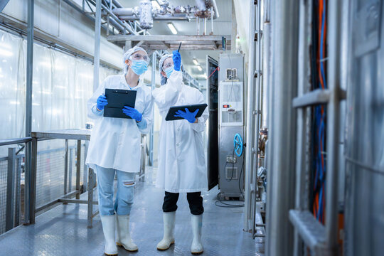 Female quality control inspectors with clipboard and tablet pointing at factory machinery, industrial plant operators checking production equipment with digital tablet and checklist