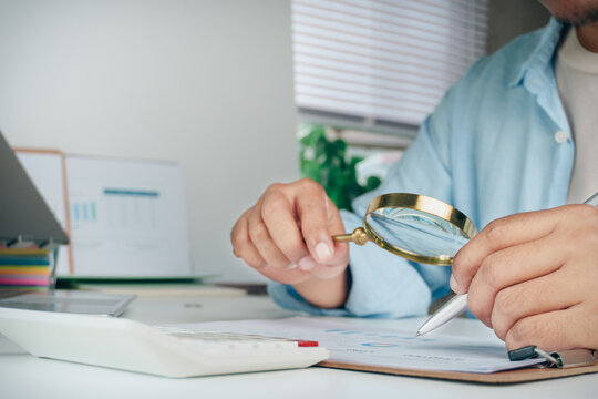 Business professional holding magnifying glass inspecting financial report paperwork with calculator on office desk, concept of data audit, analysis, auditing, and business inspection.