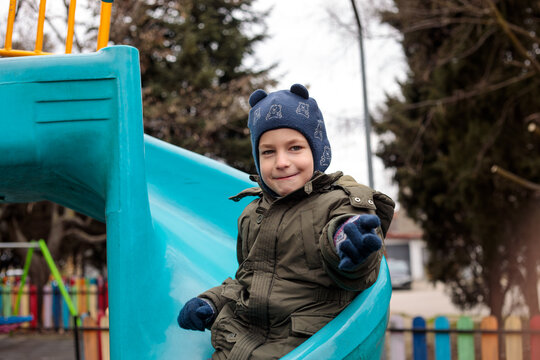 Cheerful little boy enjoys playtime at a colorful fenced playground, laughing on a round nest swing and sliding down a bright tube slide outdoors