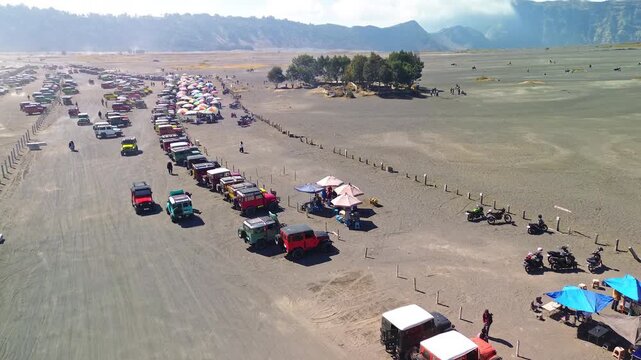 Drone view over the busy Mount Bromo jeep parking area in the Sea of Sand, showing hundreds of tour jeeps, vendor stalls, tourists and horseback riders in the Tengger caldera, Indonesia