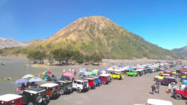 Drone flight over the busy Bromo jeep parking area and Sea of Sand, passing tourists, horseback riders and vendor stalls while approaching Mount Batok, Indonesia